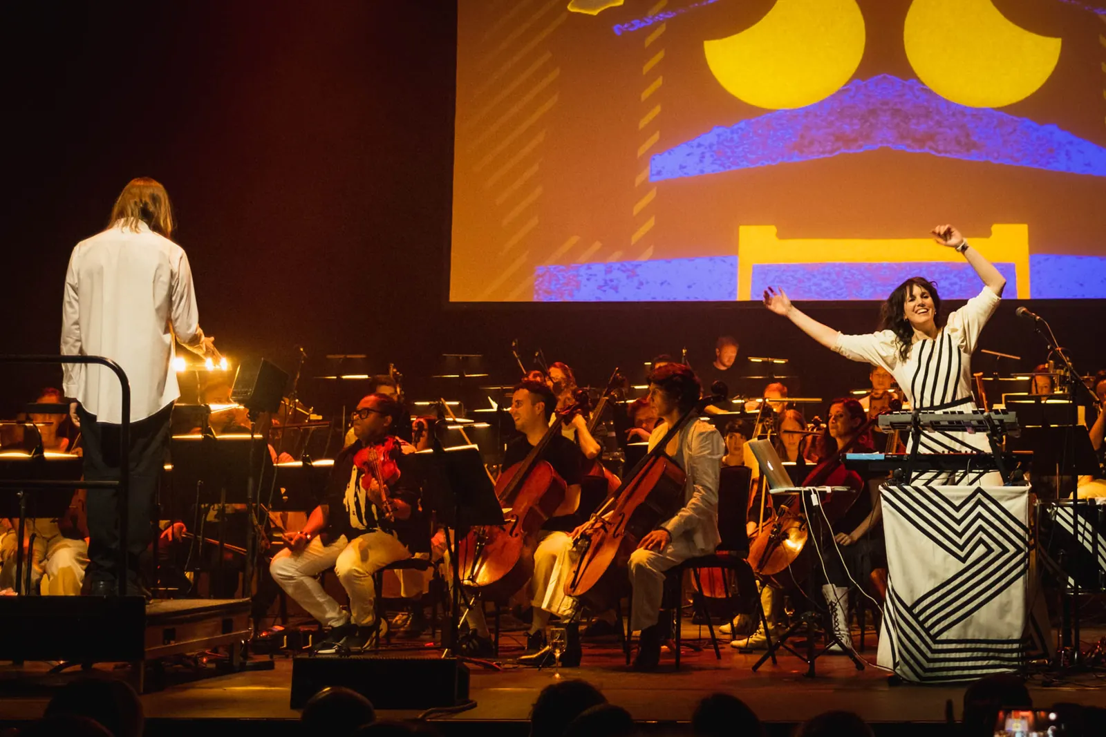 Anna Meredith raising both arms behind electronic equipment, beside seated orchestral musicians on a warmly lit stage