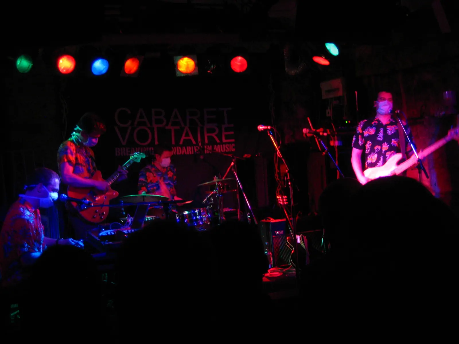 Clinic’s masked musicians in patterned shirts playing guitar, bass, drums and keyboards under coloured stage lights