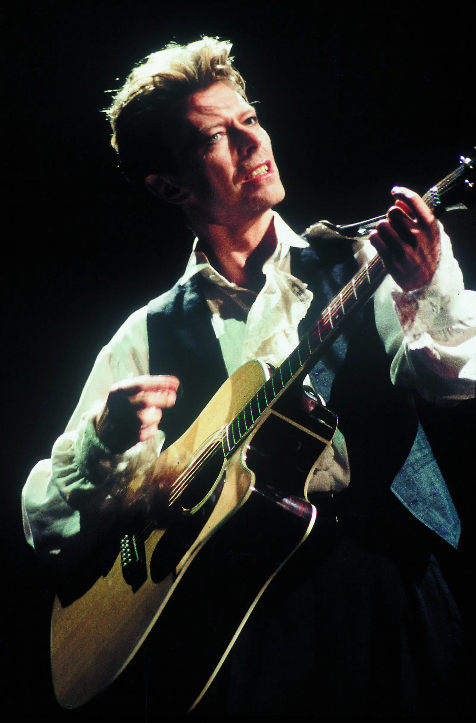 David Bowie playing an acoustic guitar in a white shirt and dark waistcoat against a dark stage background