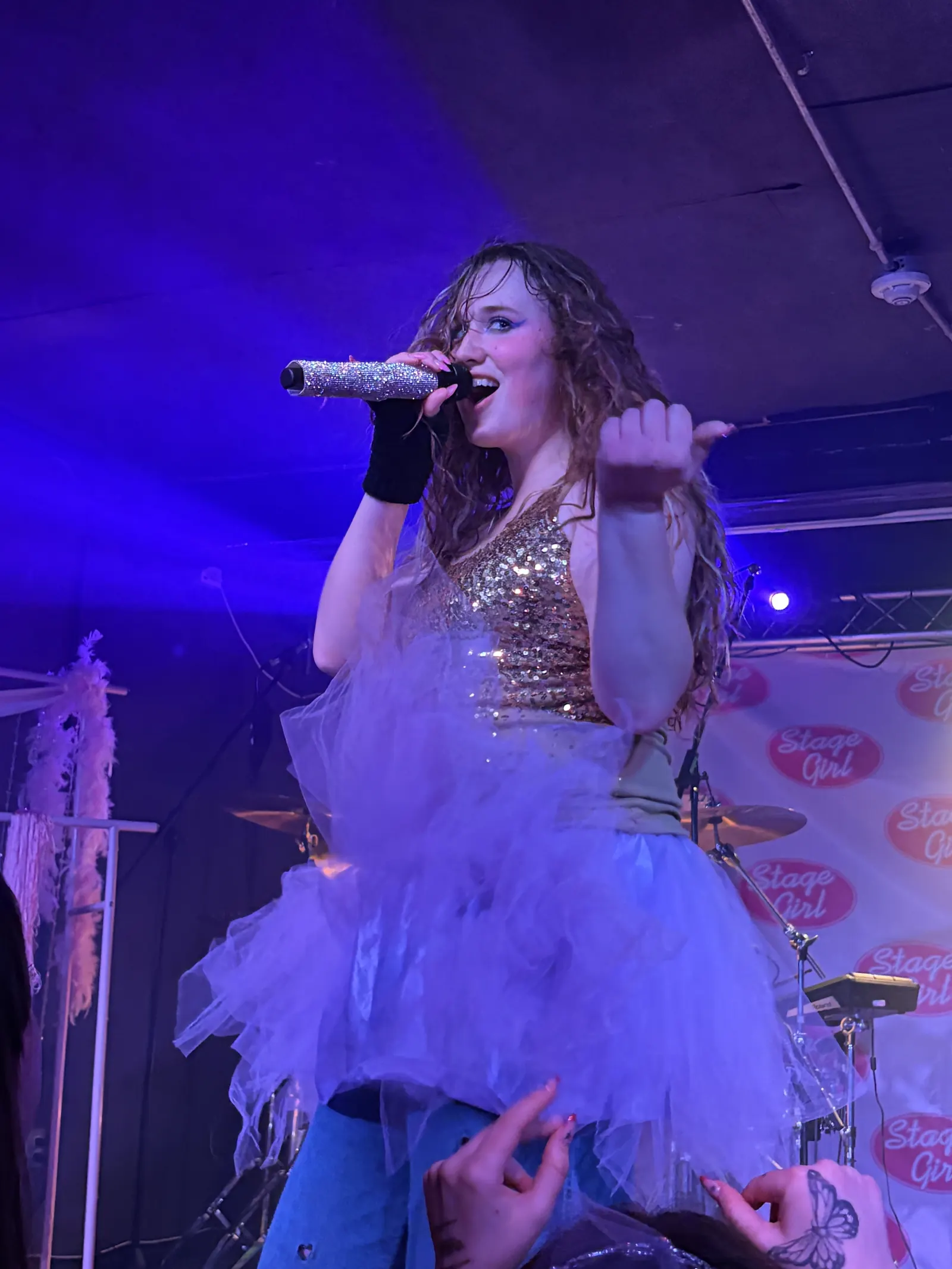 Eli singing into a sparkling microphone in a sequined top, with curly hair under purple stage lighting