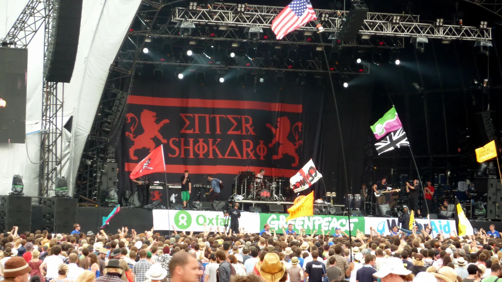 Enter Shikari performing beneath a large red-and-black band banner, with audience flags and a crowd in front of the stage