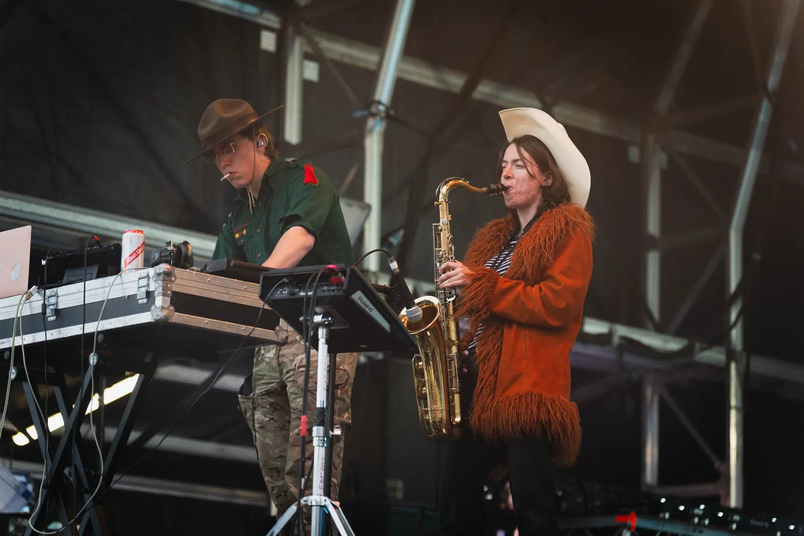 Two Fat Dog musicians onstage in 2025, one at electronic equipment and one playing saxophone in a white cowboy hat and orange fringed coat