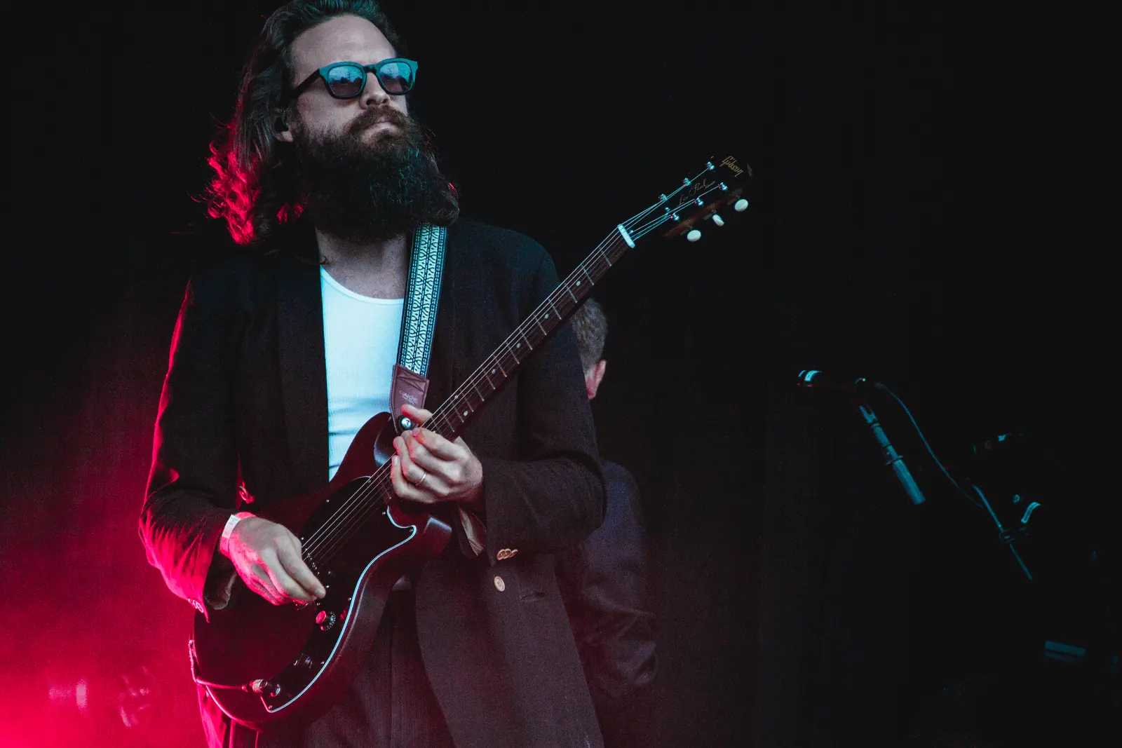 Father John Misty wearing sunglasses and a dark jacket while playing a red electric guitar under red stage lighting in 2019
