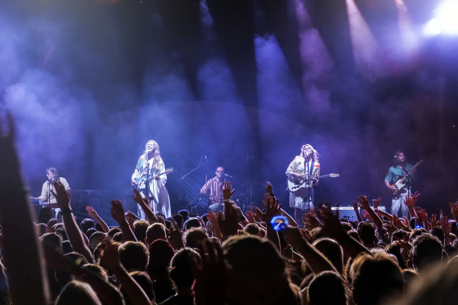 First Aid Kit singing and playing guitars with a backing band under purple stage lights, with audience members raising their hands in the foreground