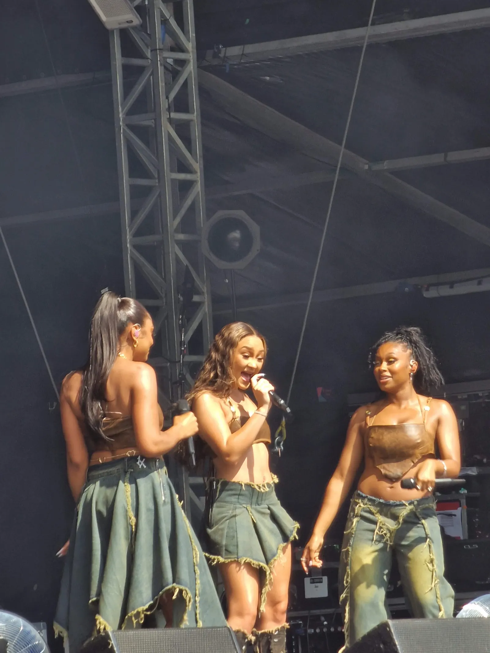 FLO’s three performers holding microphones on stage, wearing brown tops and green outfits at Mighty Hoopla