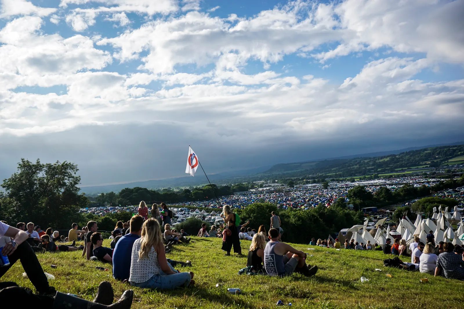 Festivalgoers sitting and standing on a grassy hillside above rows of tents under a partly cloudy blue sky