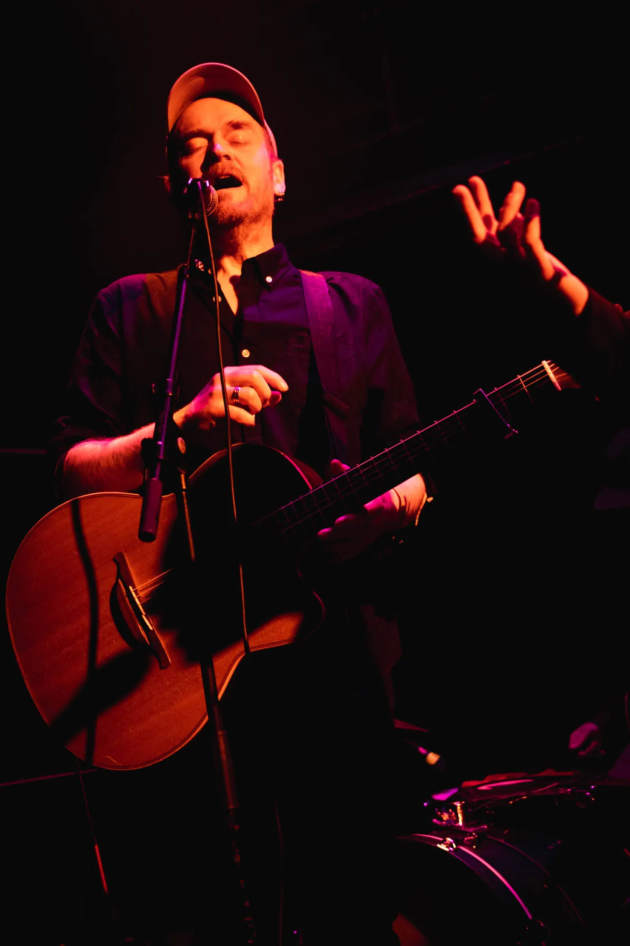 James Yorkston singing into a microphone with an acoustic guitar, wearing a cap under red and purple stage lights in 2023