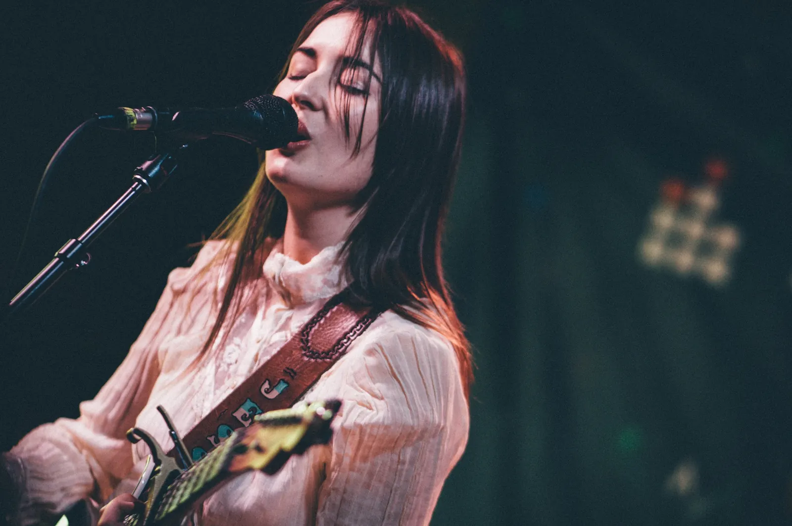 Jess Williamson singing into a microphone with a guitar, wearing a pale blouse against a dark stage background