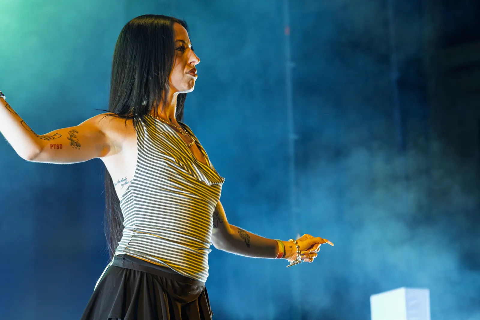 Juicy BAE with long dark hair and a striped sleeveless top, arms extended against blue stage lighting