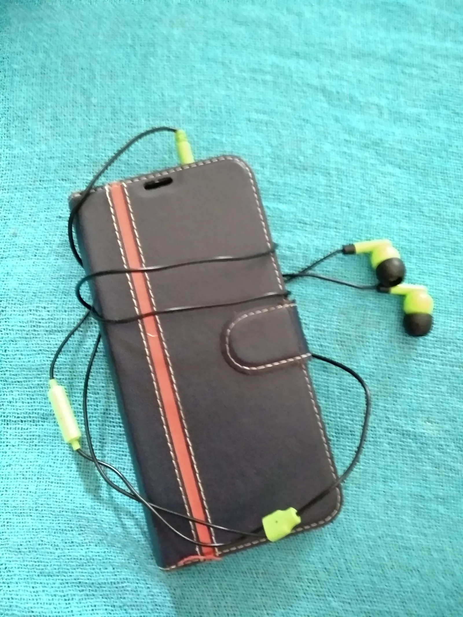A smartphone in a closed dark flip case with lime-green wired earphones on turquoise fabric
