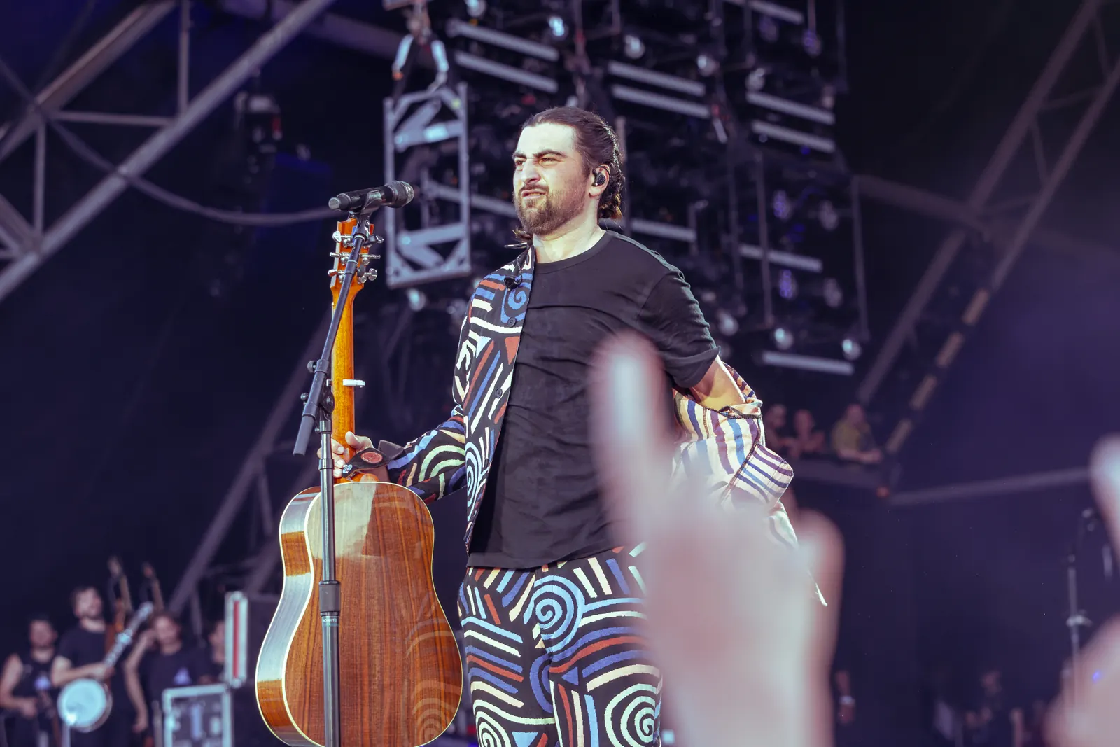 Noah Kahan beside a microphone holding an acoustic guitar at Glastonbury in 2025, wearing a black shirt and patterned trousers