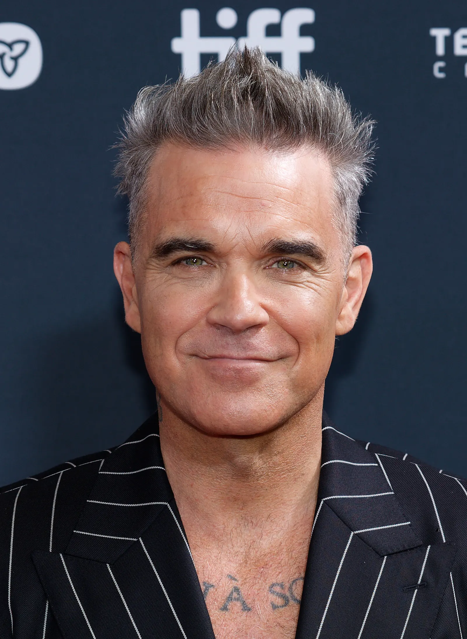 Robbie Williams with grey spiked hair and a dark pinstriped jacket in front of a TIFF backdrop